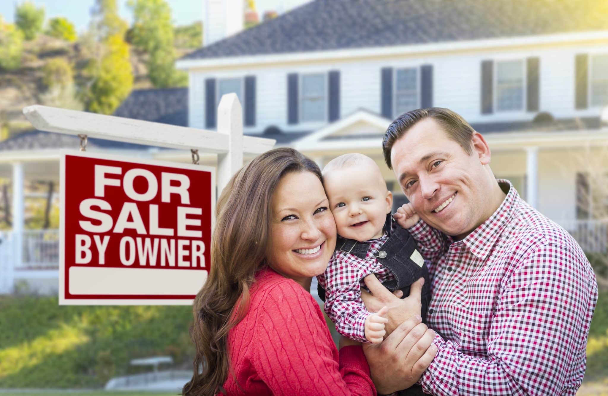 Happy family standing in front of home with For Sale By Owner sign
